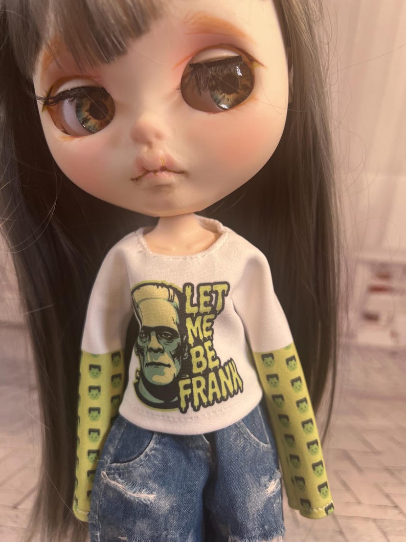 Op de afbeelding: Een pop met lang grijs haar en grote ogen draagt een wit shirt met lange mouwen met de tekst "LET ME BE FRANK" en een afbeelding van Frankenstein's monster. De mouwen zijn groen met een herhalend Frankenstein's monster ontwerp. De pop draagt ook een spijkerbroek short.