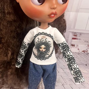 Puede incluir: Una muñeca con una camiseta blanca de manga larga con un gráfico de Medusa y pantalones vaqueros azules. Las mangas tienen un diseño en blanco y negro. La muñeca tiene cabello castaño, ojos grandes y mejillas rosadas.