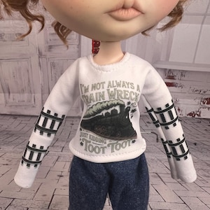 Puede incluir: Una muñeca con una camiseta blanca de manga larga con un gráfico de tren y el texto "I'm not always a train wreck, just kidding, Toot Toot". Las mangas tienen un diseño de vía de tren negra. La muñeca también lleva pantalones vaqueros azules.