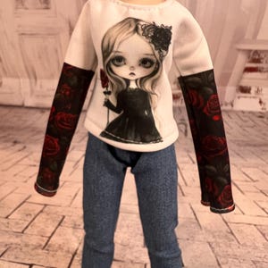 May include: A doll wearing a white long-sleeved shirt with a graphic of a girl holding a rose. The sleeves are black with red roses. The doll is also wearing blue jeans.