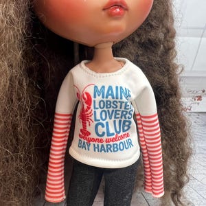 May include: A doll with long brown curly hair wearing a white long-sleeved shirt with red and white striped sleeves. The shirt has the text "MAINE LOBSTER LOVERS CLUB everyone welcome BAY HARBOUR" and is paired with black denim pants.