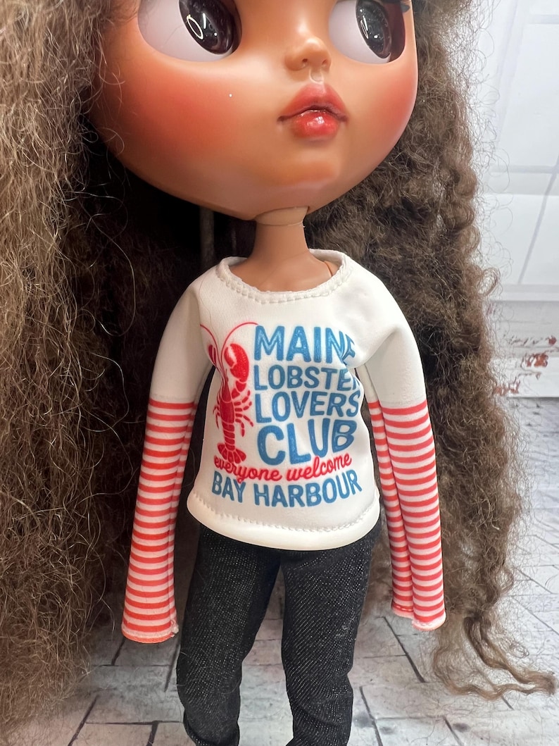 May include: A doll with long brown curly hair wearing a white long-sleeved shirt with red and white striped sleeves. The shirt has the text "MAINE LOBSTER LOVERS CLUB everyone welcome BAY HARBOUR" and is paired with black denim pants.