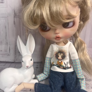 Puede incluir: Una muñeca con cabello rubio y una camisa blanca con un gráfico de una niña y conejos, combinada con jeans azules. Una figura de conejo blanco está al lado de la muñeca. La muñeca tiene ojos grandes y mejillas rosadas.