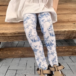 Puede incluir: Muñeca blanca con un vestido blanco y medias de estampado azul y blanco con un diseño de conejo. La muñeca lleva zapatos marrones con hebillas.