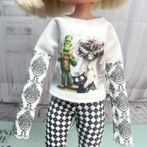 May include: A doll wearing a long-sleeved white shirt featuring a graphic print of Frankenstein's monster, his bride, and a black cat. The sleeves have a grey damask pattern. The doll also wears black and white diamond-patterned leggings.