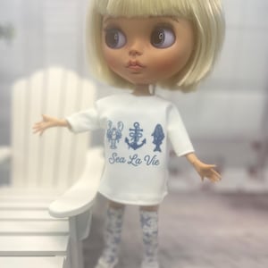 Puede incluir: Una muñeca de cabello rubio y ojos grandes que viste una camiseta blanca de manga corta con gráficos azules de una langosta, un ancla y un pez sobre el texto Sea La Vie. La muñeca lleva calcetines estampados y zapatillas blancas.