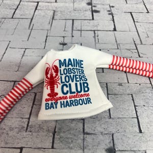 May include: White long-sleeved shirt with red and white striped sleeves. The shirt features a red lobster graphic and the text "MAINE LOBSTER LOVERS CLUB everyone welcome BAY HARBOUR" in blue and red.
