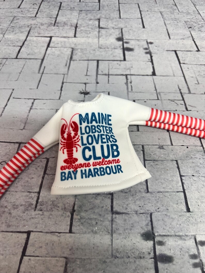 May include: White long-sleeved shirt with red and white striped sleeves. The shirt features a red lobster graphic and the text "MAINE LOBSTER LOVERS CLUB everyone welcome BAY HARBOUR" in blue and red.