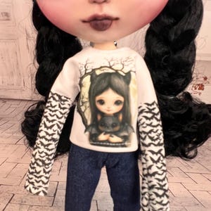 Puede incluir: Una muñeca vestida con una camiseta blanca de manga larga con un gráfico de una niña y un murciélago, y vaqueros azul oscuro. Las mangas tienen un estampado de murciélagos negros. La muñeca tiene el pelo negro trenzado y maquillaje oscuro.