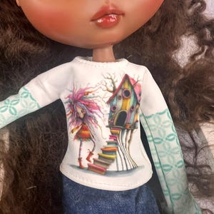 Puede incluir: Una muñeca con cabello castaño y ojos grandes que lleva una camiseta blanca de manga larga con una ilustración colorida. La ilustración presenta una niña caprichosa, una casa en el árbol y una pila de libros. Las mangas tienen un patrón verde claro y la muñeca lleva mezclilla azul.