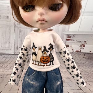 May include: A doll wearing a white long-sleeved shirt with a Halloween-themed graphic and blue denim jeans. The shirt features cartoon characters, including a penguin, ghost, and pumpkin. The sleeves are decorated with black spiders.