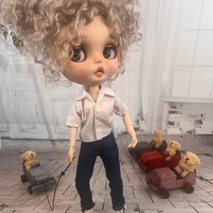 Puede incluir: Una muñeca con cabello rubio rizado, vestida con una camisa blanca, jeans azul oscuro y botas blancas. La muñeca sostiene una correa sujeta a un pequeño carro de madera con un animal de peluche dentro. Otro carro con animales de peluche está detrás.