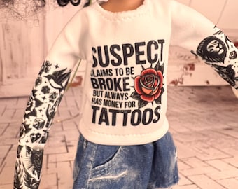 Könnte beinhalten: Eine Puppe trägt ein weißes Langarmshirt mit dem Text "SUSPECT CLAIMS TO BE BROKE BUT ALWAYS HAS MONEY FOR TATTOOS" und einer roten Rosen-Grafik. Die Puppe trägt auch zerrissene blaue Jeans. Die Arme der Puppe haben schwarz-weiße Tattoo-Ärmel.