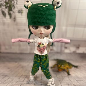 May include: A doll wearing a green crochet frog hat with white eyes, a white t-shirt with a frog graphic, green patterned pants, and white sneakers. A small frog figurine is in the background.