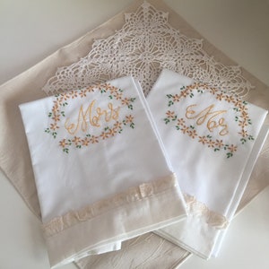 May include: Two white embroidered hand towels with a floral design and the word "Mom" in gold lettering. The towels have a lace trim and are folded on a beige surface.