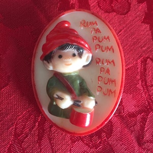 May include: A white oval-shaped Christmas bauble with a red rim. The bauble features a cartoon elf wearing a red hat and green clothing, playing a red drum. The text "RUM PA PUM PUM RUM PA PUM PUM" is printed on the bauble.