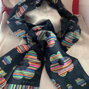 May include: A black scarf featuring a repeating pattern of multi-coloured striped flowers in shades of blue, green, yellow, pink, and orange. The scarf has a striped border along the bottom edge and the word ECHO is printed in white near the corner.