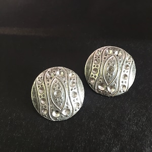 May include: A pair of silver earrings with a circular design featuring a textured pattern of raised dots and lines.