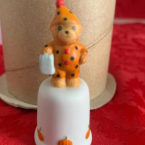 May include: A ceramic thimble featuring a bear wearing an orange and black polka-dotted outfit and hat, holding a small white item. The thimble is white with orange pumpkin designs. A festive, seasonal collectible.