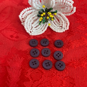 May include: A handcrafted flower ornament made of white seed beads with a centre of yellow and green beads, accompanied by eight small, round, dark purple buttons, each featuring four holes arranged in a square pattern on the surface.