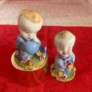 Peut inclure: Deux figurines en céramique représentant des enfants aux cheveux clairs vêtus de tenues bleues. Un enfant est assis sur une base cylindrique dorée entouré de fleurs et d'une coccinelle, l'autre est agenouillé sur un socle herbeux avec un bouquet.