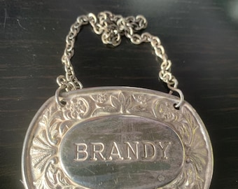 Può includere: Etichetta per bottiglia in argento con un design ornamentale e la parola "BRANDY" incisa al centro. L'etichetta è attaccata a una catena d'argento.