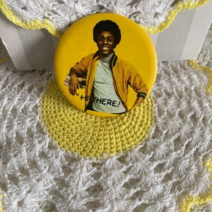 May include: A yellow button with an image of a smiling person in a yellow jacket and the words "Hi There!". The button is on a white and yellow crocheted background.