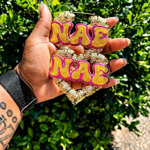 Op de afbeelding: Goudkleurige oorringen met de naam "NAE" in een bubble letter-stijl, omlijnd in roze en gevuld met gouden glitter. De oorbellen worden in een hand gehouden tegen een achtergrond van groen gebladerte.