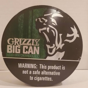 Puede incluir: Una lata redonda, verde, negra y plateada con un gráfico de un oso grizzly y el texto "GRIZZLY BIG CAN LONG CUT WINTERGREEN". La lata tiene una etiqueta de advertencia que dice "ADVERTENCIA: Este producto no es una alternativa segura a los cigarrillos."