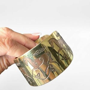 May include: A wide gold-toned metal cuff bracelet featuring an engraved repeating pattern of horses and intricate knotwork designs. The polished surface reflects light, highlighting the detailed etched outlines of the equine figures.