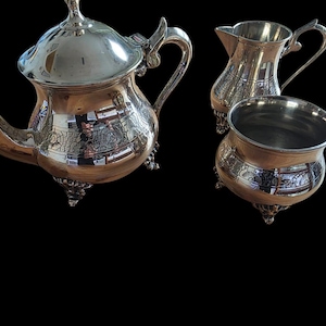 Può includere: Un servizio da tè argentato con dettagli ornati, tra cui una teiera con coperchio, una lattiera e una zuccheriera. Il set presenta piedini e manici decorativi, che riflettono la luce e creano un'estetica classica.
