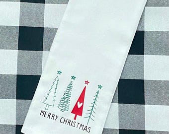 Puede incluir: Toalla de cocina blanca con un fondo de cuadros de búfalo blanco y negro. La toalla presenta una ilustración roja, verde y blanca de tres árboles de Navidad con estrellas y el texto "Feliz Navidad".