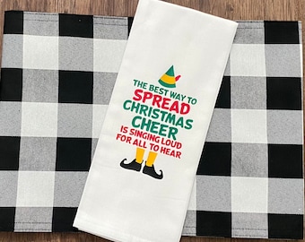 Puede incluir: Toalla de cocina blanca con un fondo a cuadros blanco y negro. La toalla presenta un duende de dibujos animados con el texto "The best way to spread Christmas cheer is singing loud for all to hear."