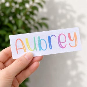 May include: A rectangular white sticker featuring the name Aubrey in a colorful, rounded, hand-lettered font. Each letter displays a gradient transition of colors, including orange, yellow, light blue, dark blue, purple, and coral pink.