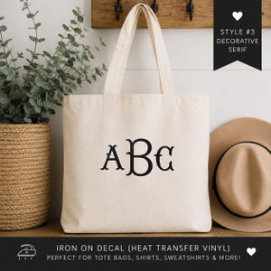 May include: A natural-colored canvas tote bag featuring a black heat transfer vinyl decal with the letters ABC in a decorative serif font. The bag has two shoulder straps and is displayed with a tan hat and a green plant in a woven basket.