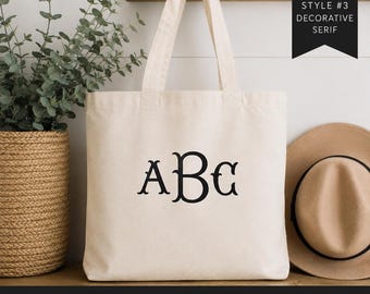May include: A natural-colored canvas tote bag featuring a black heat transfer vinyl decal with the letters ABC in a decorative serif font. The bag has two shoulder straps and is displayed with a tan hat and a green plant in a woven basket.