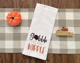 Puede incluir: Toalla de cocina blanca con el texto "Gobble 'til you Wobble" en negro y naranja. Una calabaza naranja decorativa y un letrero de bloque de madera que dice "Happy Harvest" también están en la imagen. La toalla está sobre un camino de mesa a cuadros grises y blancos.