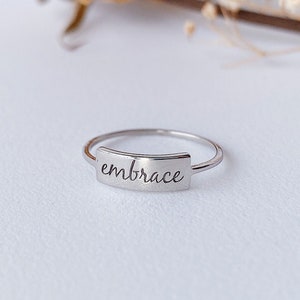 Puede incluir: Un anillo de plata con una banda rectangular que dice "embrace" en escritura cursiva.