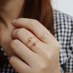 Puede incluir: Tres anillos de oro en un dedo. Los anillos son finos y delicados. Un anillo tiene un pequeño diseño de hoja, otro tiene una pequeña perla y el tercer anillo tiene la letra "W" en él.