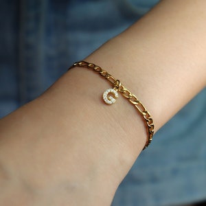 May include: A gold chain bracelet with a charm in the shape of the letter G, encrusted with small clear stones.