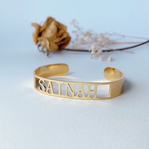 May include: Gold-toned metal bracelet with the name "SAINAH" cut out in the metal.
