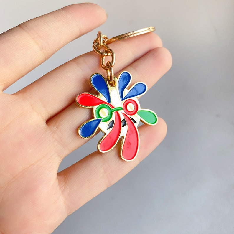 May include: Gold-coloured keychain with a colourful abstract design. The design features red, blue, green, and white paint-splatter shapes.