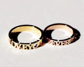 May include: Two gold rings with the words "LOVE YOU" and "FOREVER" engraved on them. The rings are on a white surface.
