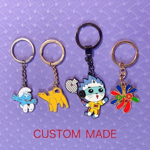 May include: Four keychains featuring different cartoon characters. The keychains are gold and silver with blue, red, yellow, and green accents. The characters include a blue Smurf, a yellow camel, a white cat with a blue mask, and a red, yellow, and green abstract design.