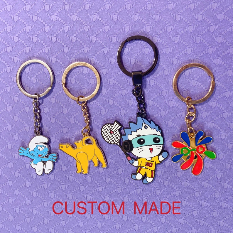 May include: Four keychains featuring different cartoon characters. The keychains are gold and silver with blue, red, yellow, and green accents. The characters include a blue Smurf, a yellow camel, a white cat with a blue mask, and a red, yellow, and green abstract design.