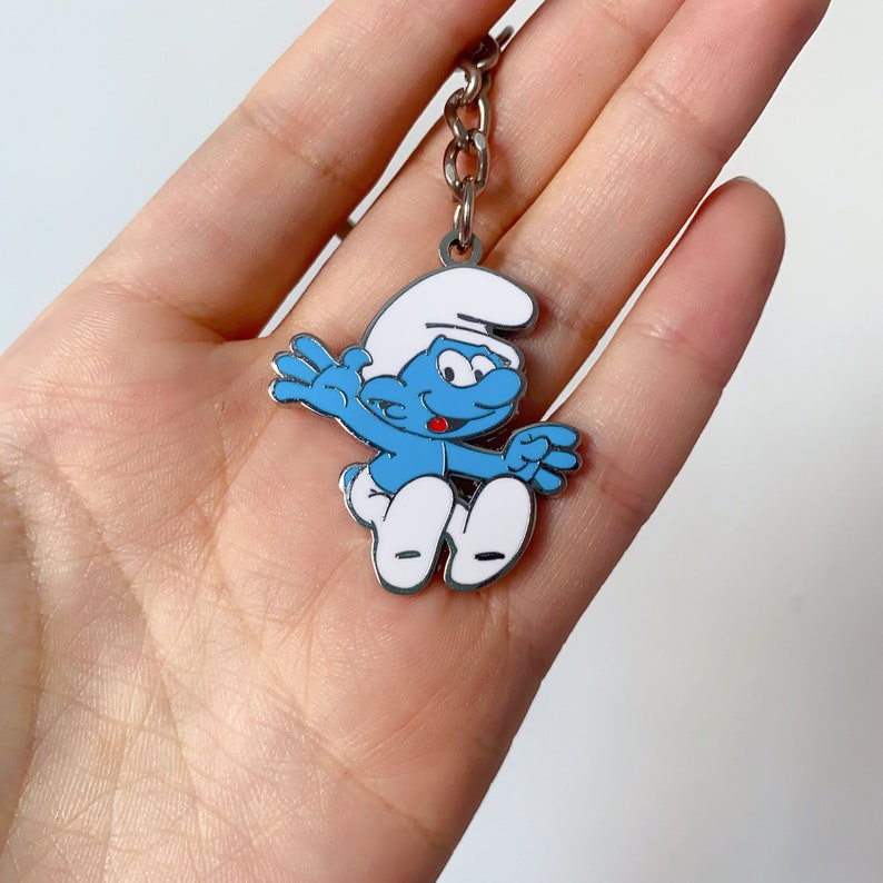 May include: A silver keyring featuring a blue Smurf character with a white hat and a red mouth. The Smurf is depicted in a jumping pose with arms outstretched.