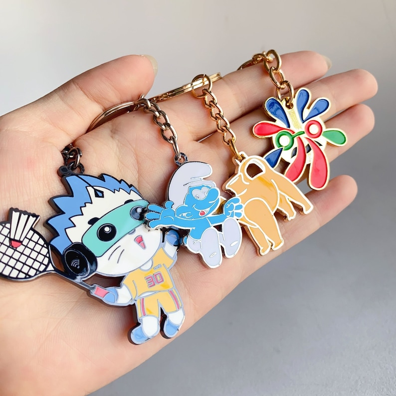 May include: A set of five colourful keychains featuring cartoon characters. The keychains are made of metal and have a shiny finish. The characters include a blue Smurf, a yellow character with a red and blue design, a white and blue character with a red and blue design, a blue and white character with a red and blue design, and a white and blue character with a red and blue design.