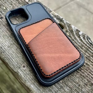 Puede incluir: Una funda de teléfono negra con un porta tarjetas de cuero marrón unido a la parte posterior. El porta tarjetas tiene un borde cosido.