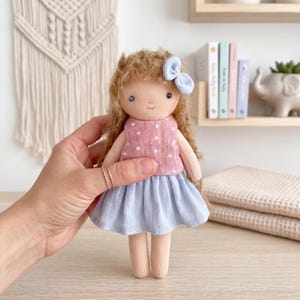 Op de afbeelding: Een stoffen pop met krullend bruin haar en een lichtblauwe strik. De pop draagt een roze mouwloos topje met witte stippen en een effen lichtblauwe rok. De pop heeft eenvoudige geborduurde gelaatstrekken en ledematen van lichte stof.