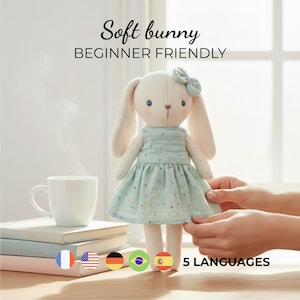Puede incluir: Una muñeca de conejito suave y blanco con orejas largas y un vestido azul claro. La muñeca tiene un lazo en la cabeza y la sostiene una persona. El texto "Soft bunny BEGINNER FRIENDLY" está encima de la muñeca. Una taza blanca y libros están en el fondo.